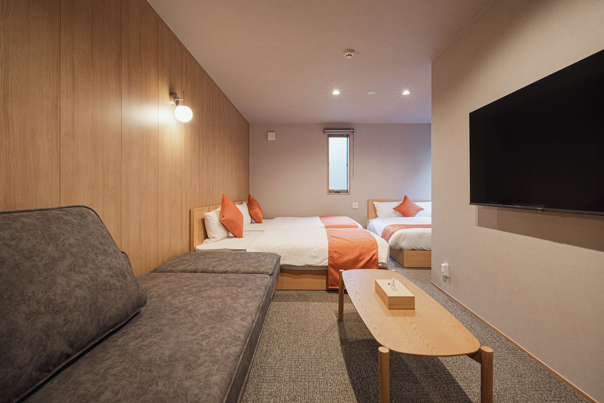 A modern hotel room with a sofa, two beds with orange pillows, a TV, and a window.