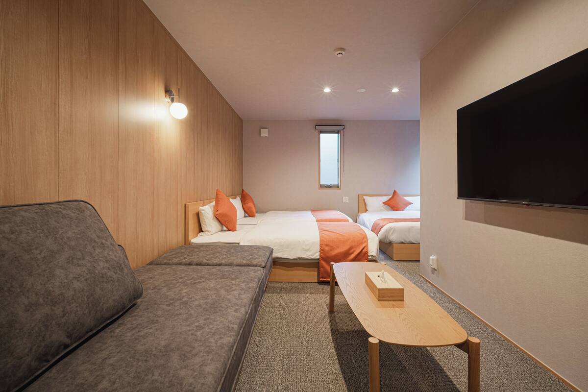 A modern hotel room with a sofa, two beds with orange pillows, a TV, and a window.