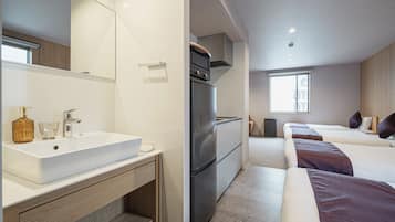 A modern hotel room with a large bathroom sink, a built-in oven, a refrigerator, and two beds with dark bedding.