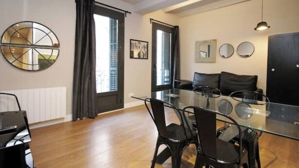 A modern living room with a glass dining table, black chairs, a black sofa, and a large window with curtains. - Spacious 2BR, quick access to Downtown (Barcelona)