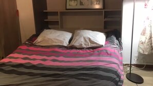 Uma cama com colcha estampada, uma luminária de cabeceira e uma prateleira na parede com quadros.