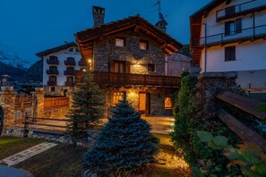 Un bâtiment en pierre doté d’un balcon en bois, une enseigne indiquant « HÔTEL BERTHO », et une chaîne de montagnes en arrière-plan.