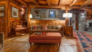 Interieur van een blokhut met houten plafond, stenen muren, een bank, een stoel, een tafel en een open haard.