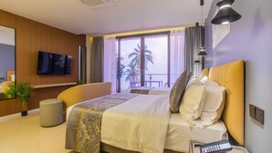 Quarto de hotel moderno com uma cama grande, televisão de tela plana, mesa de cabeceira com controle remoto e vista para palmeiras pela janela.