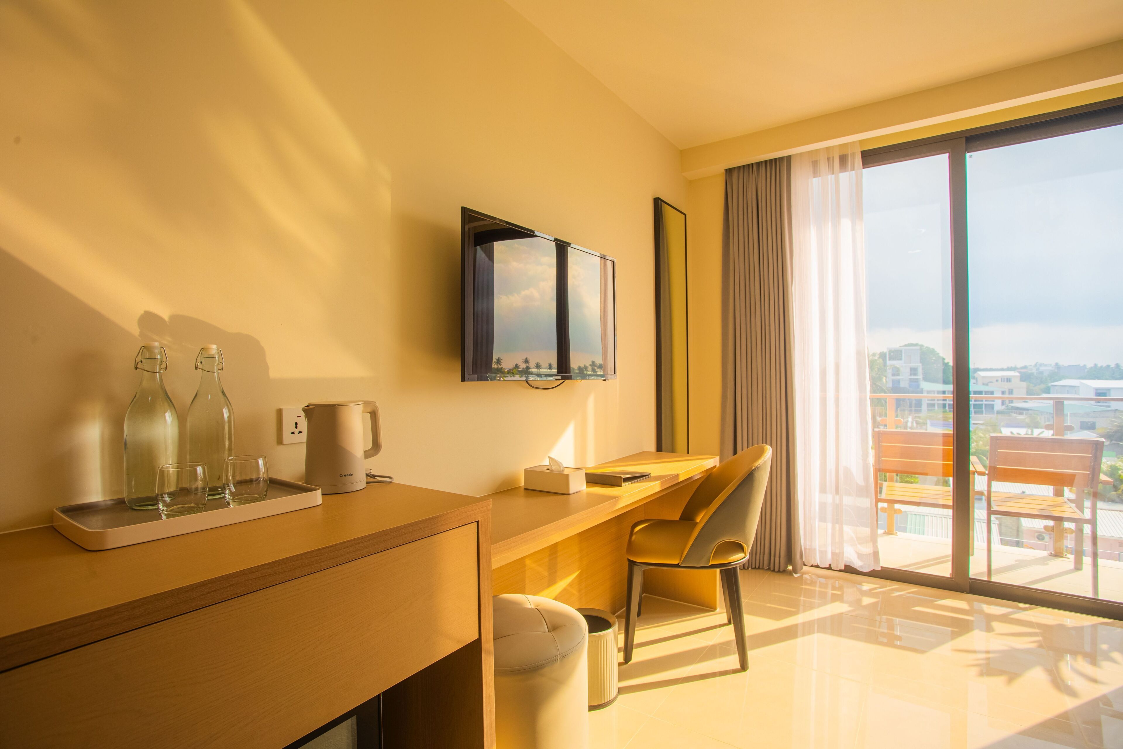 A modern hotel room with a wooden desk, a chair, a large window with a city view, and a flat-screen TV mounted on the wall.
