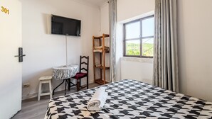 Een kamer met een bed, een aan de muur bevestigde televisie, een raam met gordijnen, een klein tafeltje met een stoel, en een houten plank.