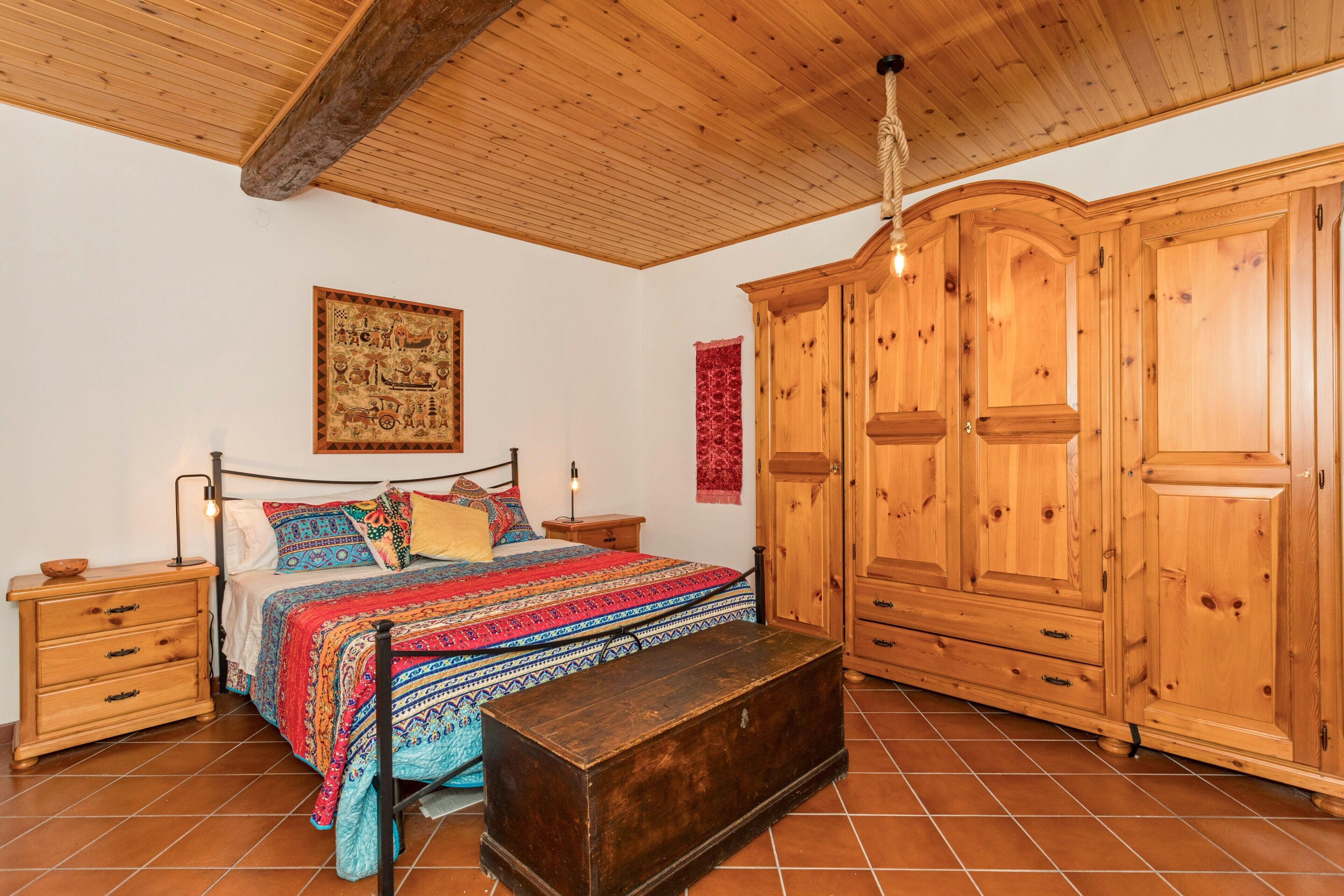 Een slaapkamer met een houten plafond, een bed met beddengoed met een patroon, een houten garderobe en een bankje.