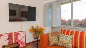 A living room with a sofa, a flat-screen TV, artwork, a potted plant, and a window with a view. - Apartment 'Casa Valerio' with Sea View, Wi-Fi and Air Conditioning (Fiumefreddo di Sicilia)
