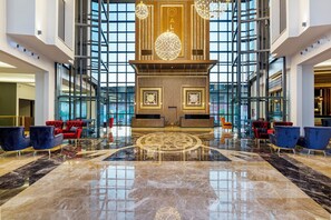 A spacious lobby with a polished marble floor, red and blue seating, and a large decorative wall with a clock.