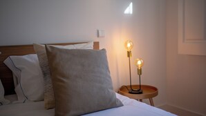 Ein ordentlich bezogenes Bett mit einem grauen und einem weißen Kissen sowie einem hölzernen Kopfteil. Neben dem Bett steht ein kleiner Nachttisch mit einer Lampe.