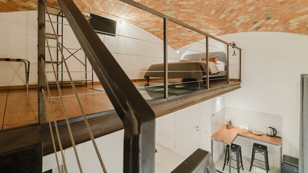 A loft-style bedroom with a brick ceiling, a glass mezzanine, and a kitchen area below. - Quadruple Room with Balcony Overlooking the Main Street and Square, Air Conditioning (Calangianus)