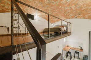 A loft-style bedroom with a brick ceiling, a glass mezzanine, and a kitchen area below. - Quadruple Room with Balcony Overlooking the Main Street and Square, Air Conditioning (Calangianus)