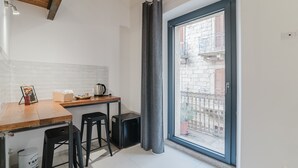 A small kitchen with a wooden table, two black stools, a black suitcase, and a window with a view of a brick building. - Quadruple Room with Balcony Overlooking the Main Street and Square, Air Conditioning (Calangianus)
