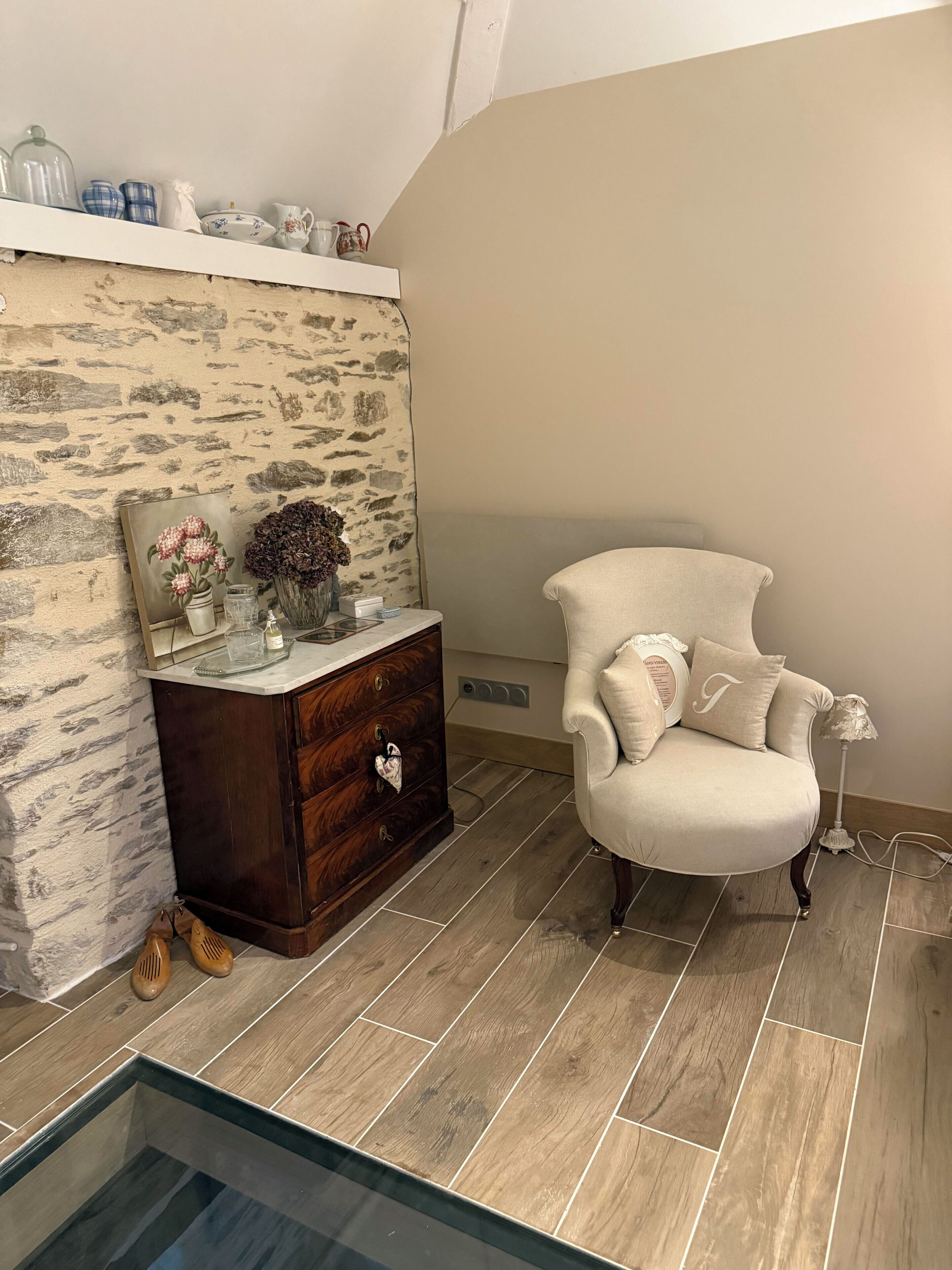 Quarto com parede de padrão em pedra, cômoda de madeira, poltrona cor creme e mesa com tampo de vidro.