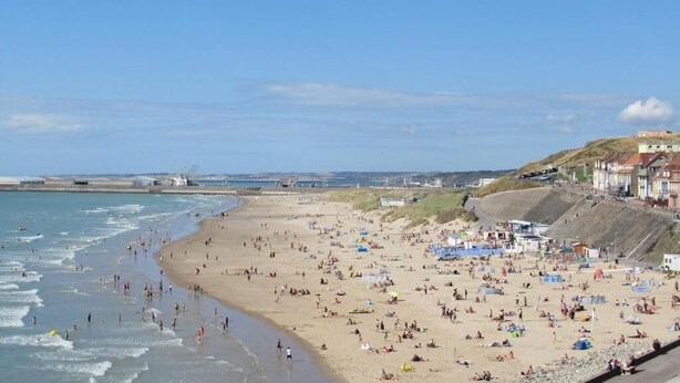 Ein überfüllter Strand mit Menschen und Sonnenschirmen, ein klarer Himmel und Gebäude im Hintergrund.