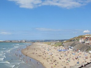 Een druk strand met mensen en parasols, een heldere lucht en gebouwen op de achtergrond.