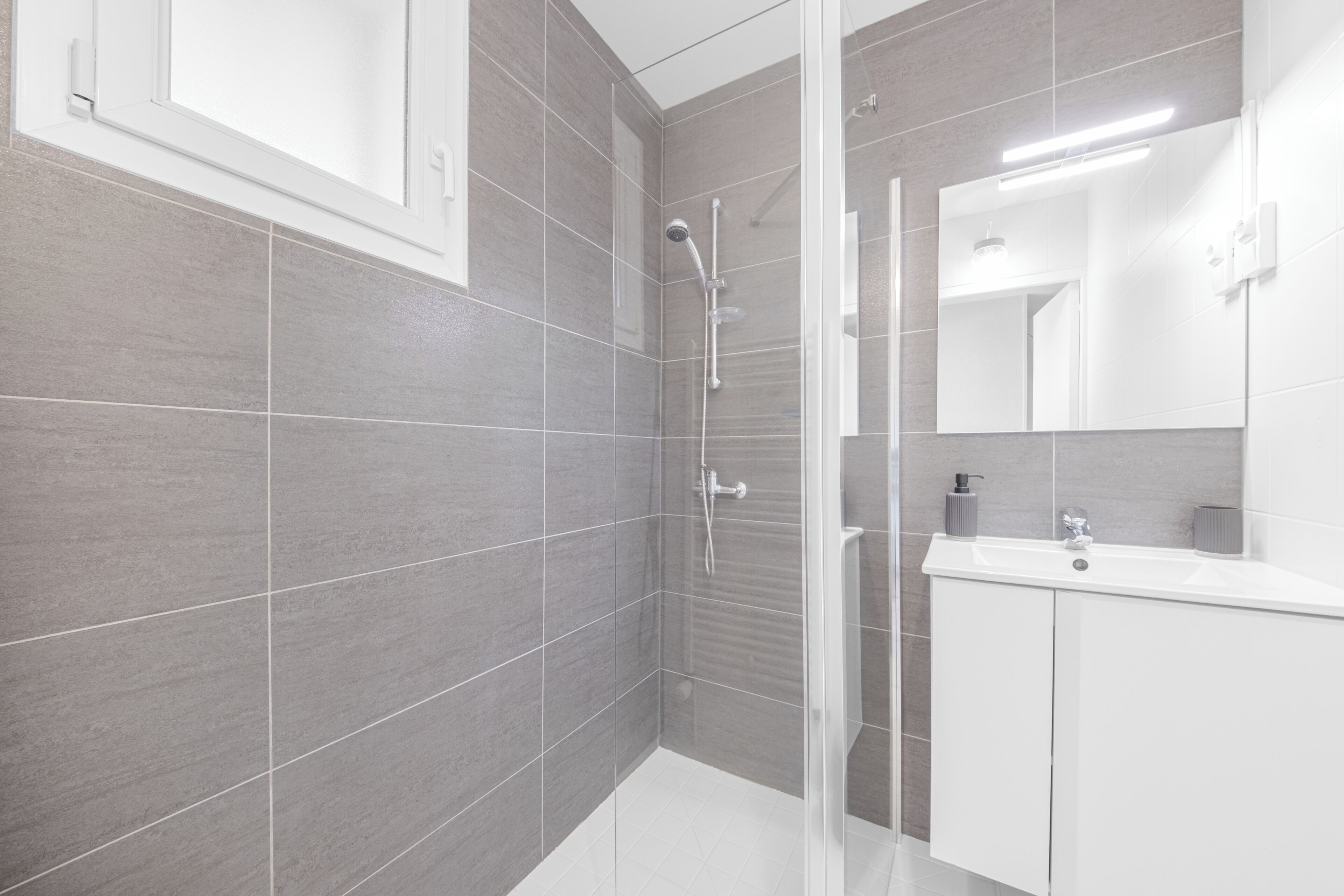 A modern bathroom with grey tiles, a glass shower enclosure, a white sink, and a mirror.
