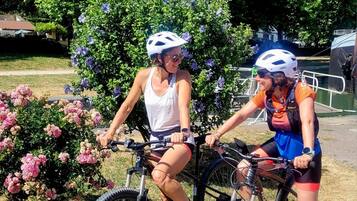 Deux vélos sont garés près d’un arbuste en fleurs, avec une zone herbeuse et des arbres en arrière-plan.