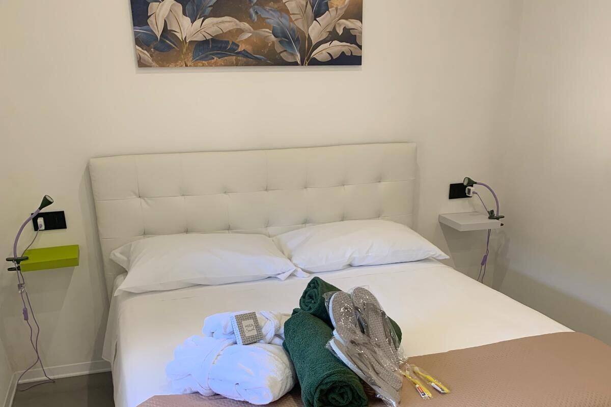 Una cama bien tendida con sábanas blancas, una toalla verde y zapatillas, una mesita de noche con una lámpara, y un cuadro en la pared.