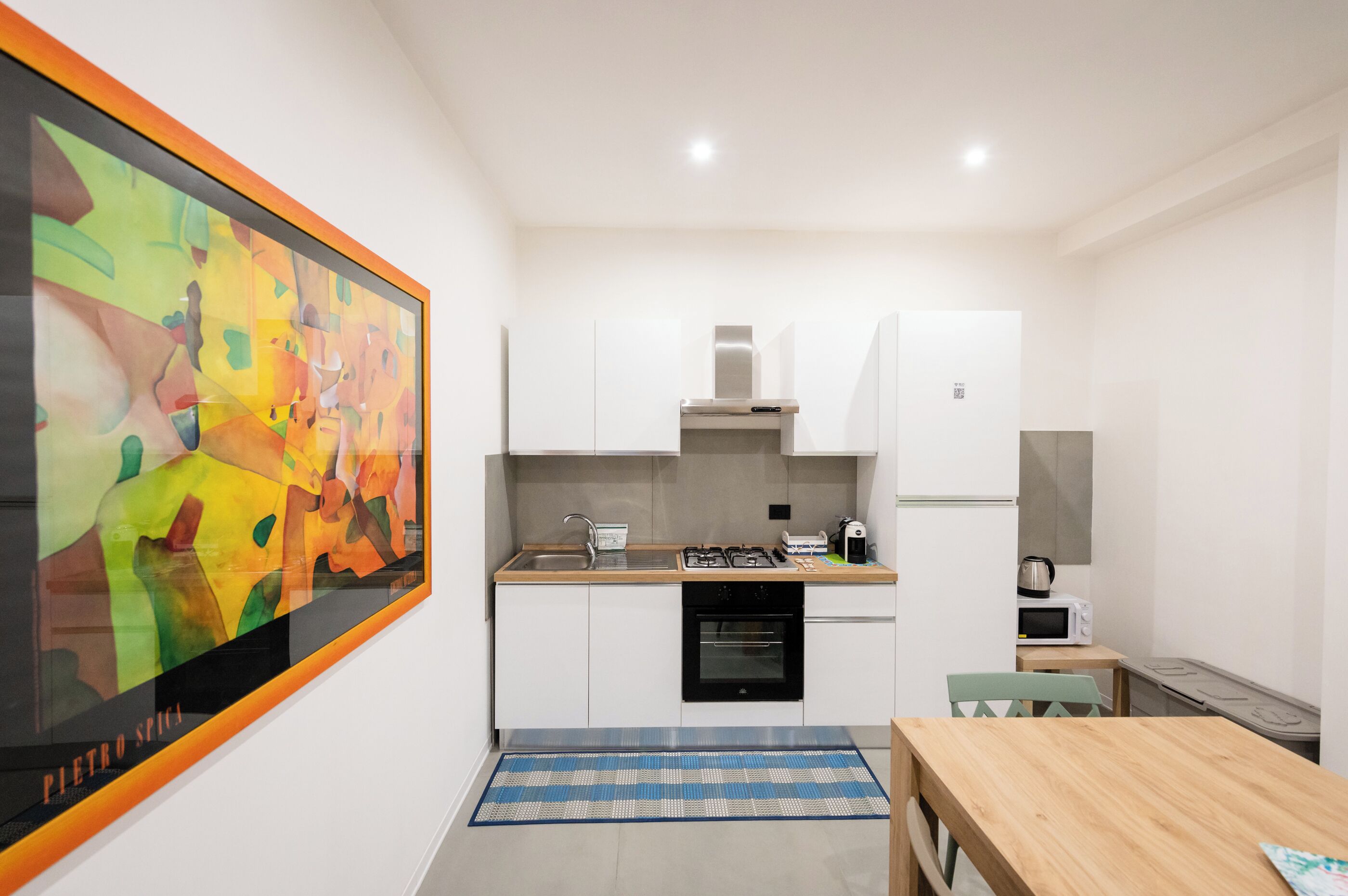 A compact kitchen with white cabinets, a stove, microwave, and a kettle. A large abstract painting adorns the wall.