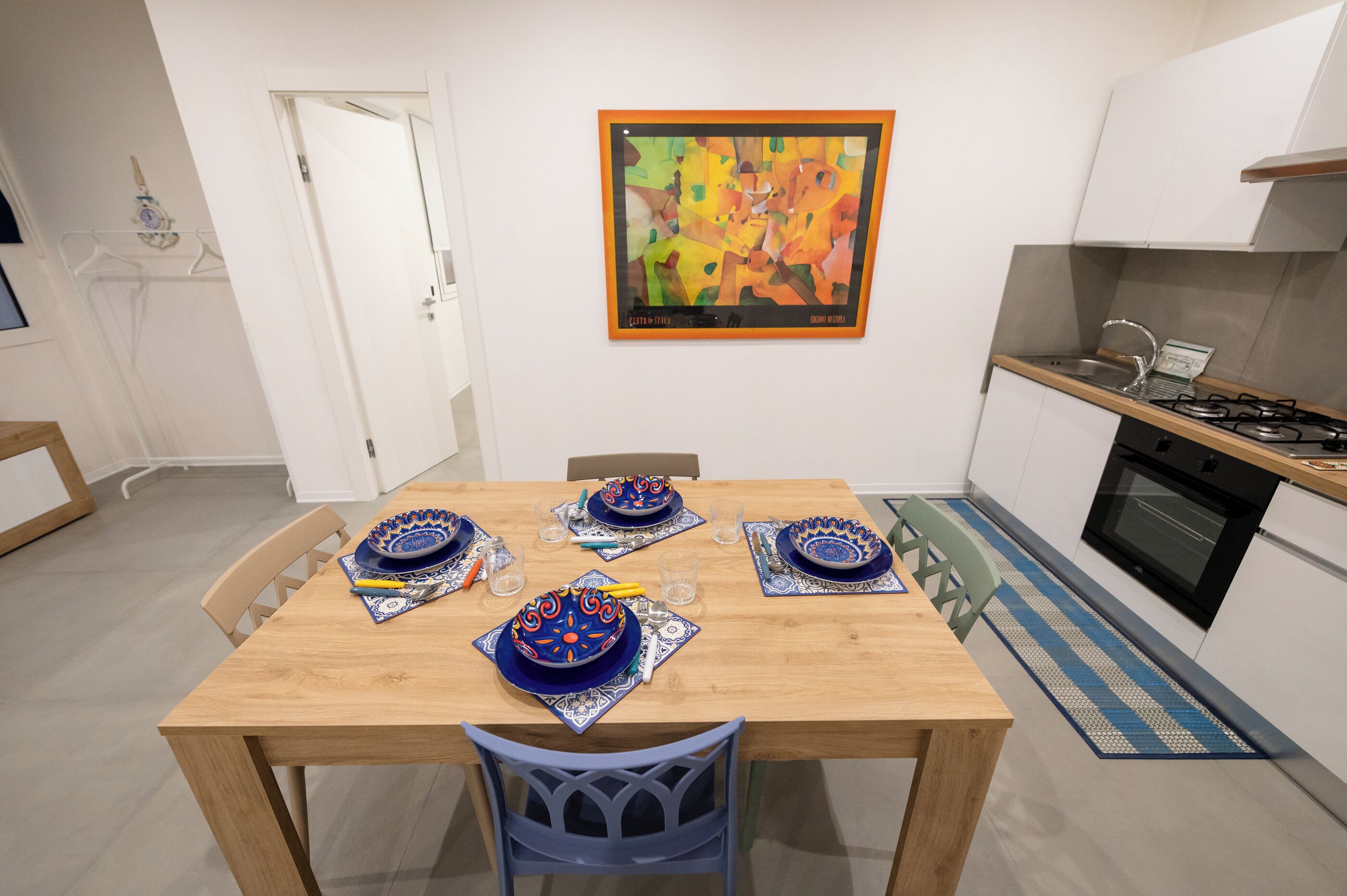 A dining area with a wooden table set for four, a kitchen with white cabinets and a stove, and a colorful abstract painting on the wall.