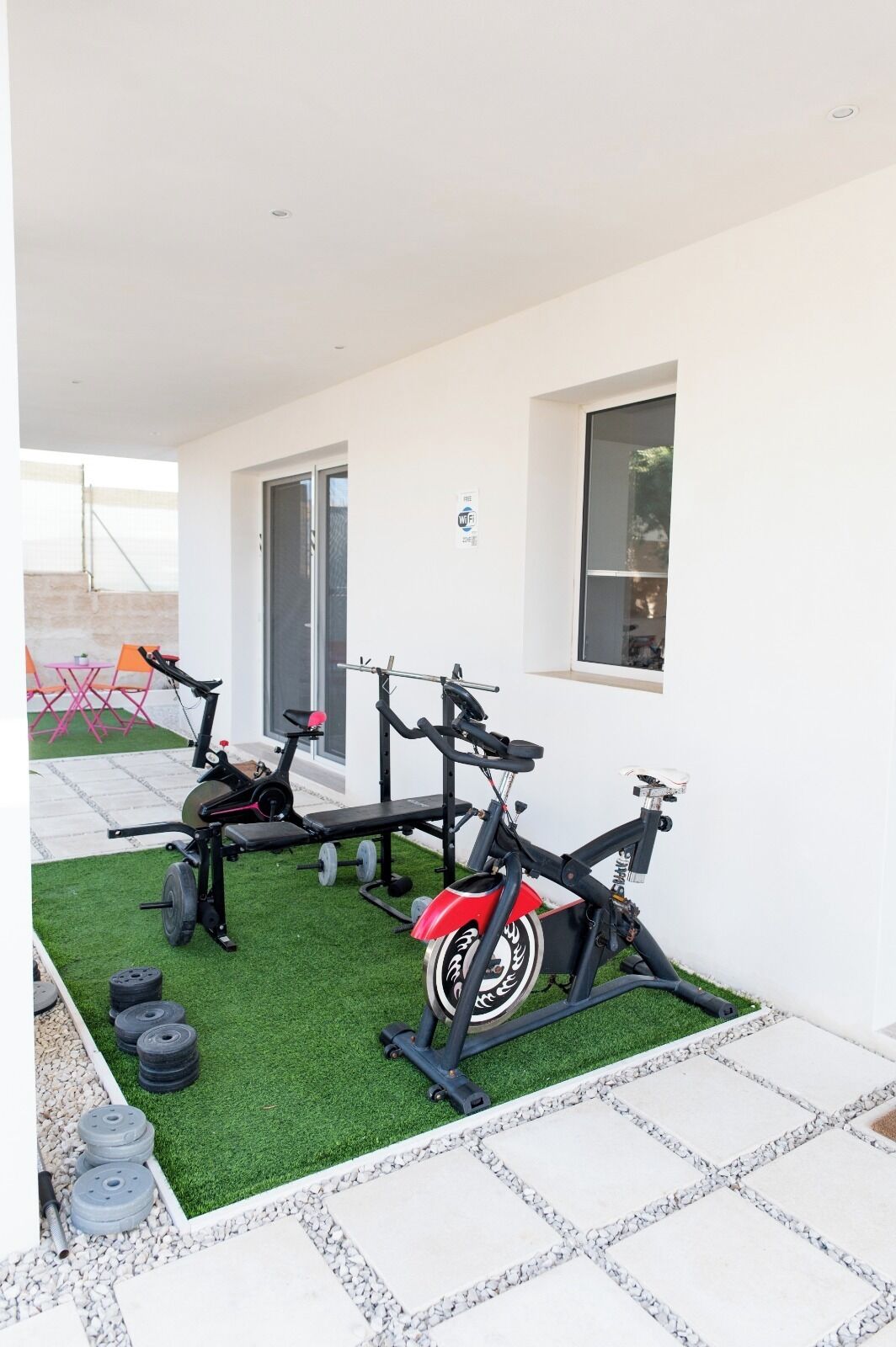 A home gym with exercise equipment, including a stationary bike, a bench press, and a weight bench, on a green artificial turf area.