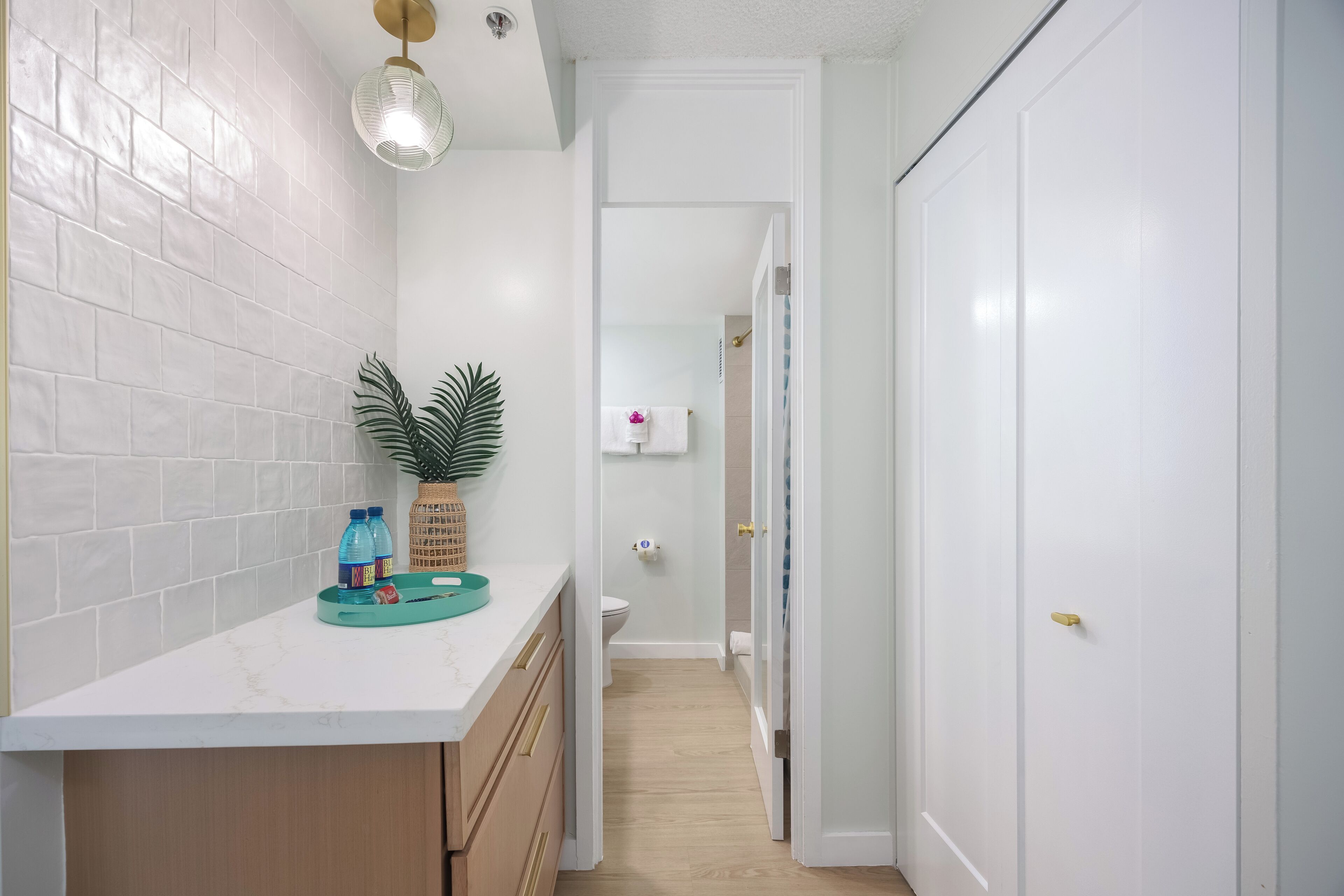 A narrow hallway with a white tiled wall, a wooden cabinet, and a countertop with a plant and water bottles.