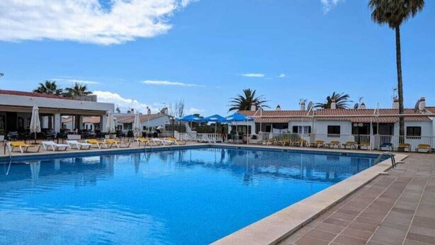 A swimming pool with lounge chairs and umbrellas, surrounded by buildings with tiled roofs and palm trees. - Casa Carlito 153 - zone 3 Son Bou (San Jaime Mediterráneo)