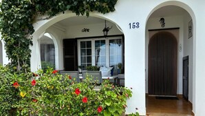 A white building with a number '153' on the wall, a covered porch with chairs, and a door with a decorative handle. - Casa Carlito 153 - zone 3 Son Bou (San Jaime Mediterráneo)