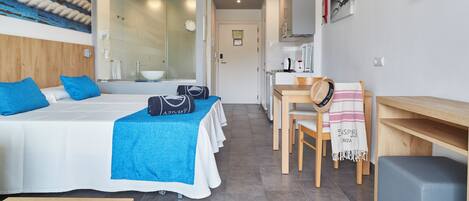 A modern hotel room with a large bed, a desk, and a view into the kitchen area.
