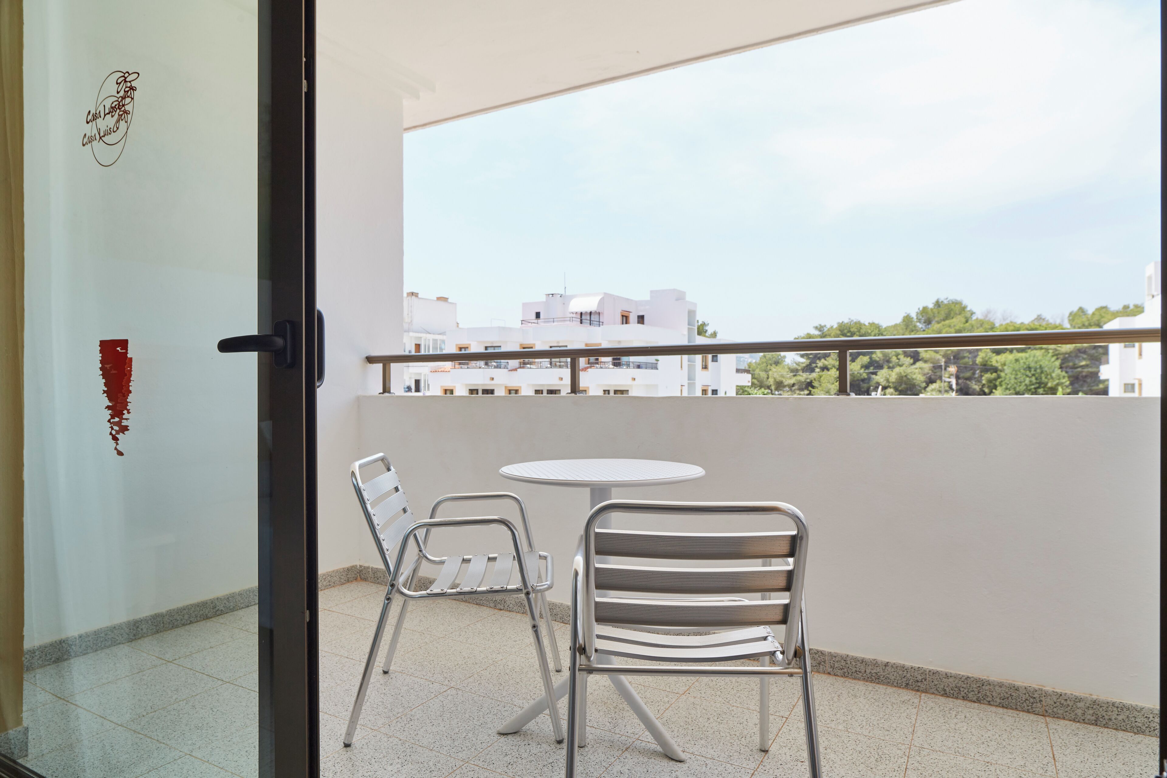 Un balcón con dos sillas de metal y una pequeña mesa redonda, con vistas a un paisaje urbano de edificios blancos y áreas verdes.