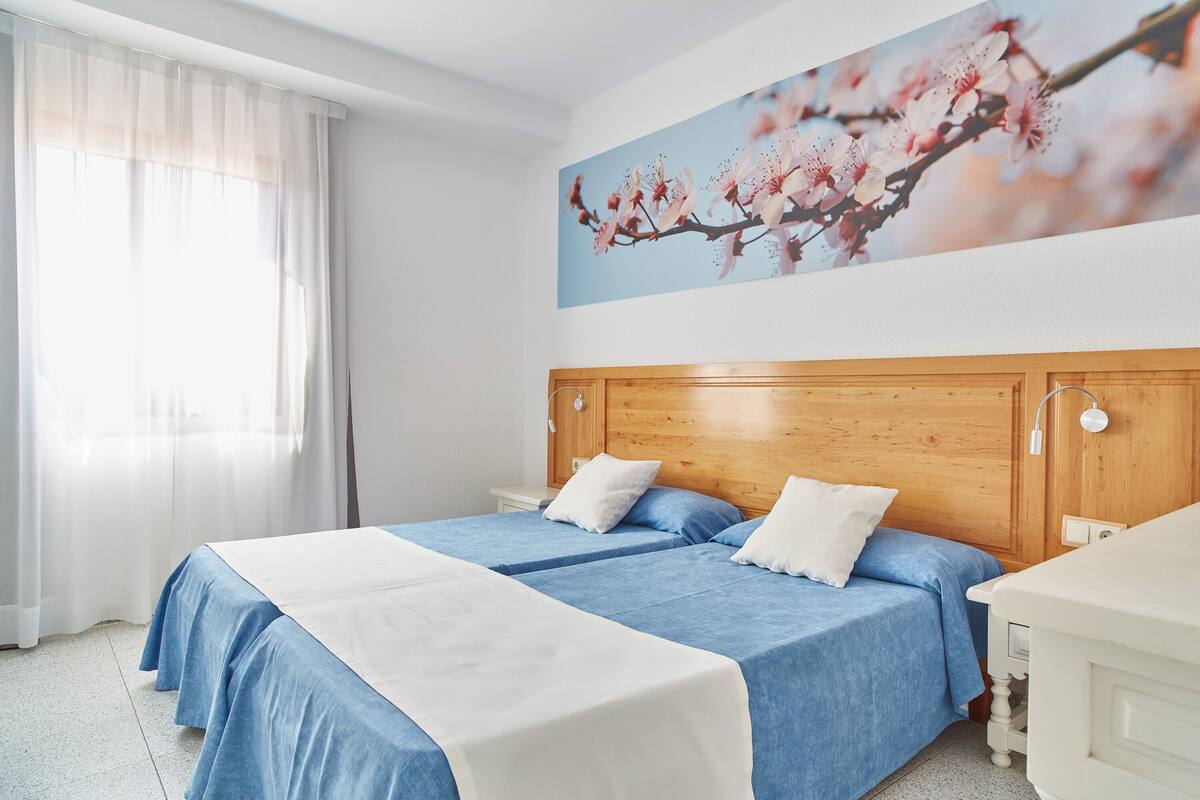 Une chambre d’hôtel avec une tête de lit en bois, deux lits avec une literie bleue et blanche, une table de chevet blanche et une lampe fixée au mur.