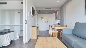 Een moderne woonkamer met een bank, een houten salontafel, een bed met een witte spreidek en een eethoek met een tafel en stoelen, en een keuken op de achtergrond.