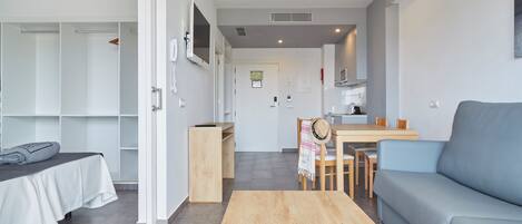 A modern living room with a sofa, a wooden coffee table, a bed with a white bedspread, a dining area with a table and chairs, and a kitchen in the background.