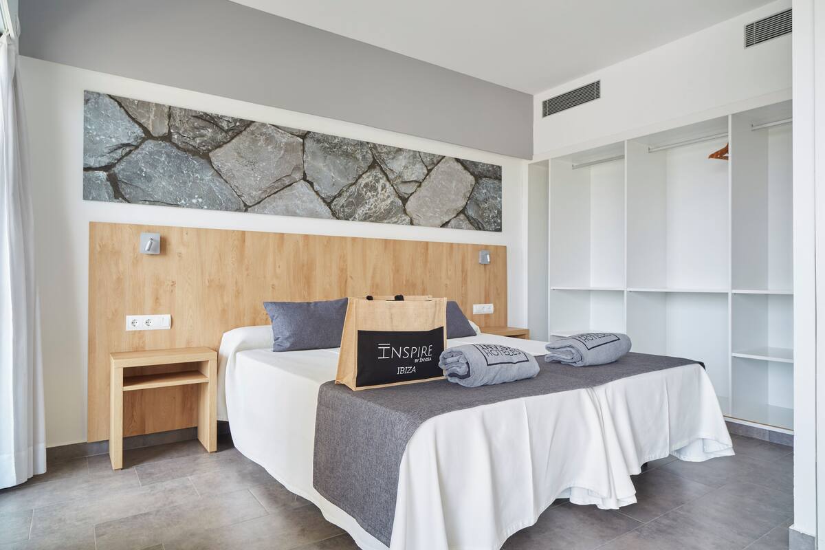 Une chambre moderne avec un mur en pierre, une tête de lit en bois, une table de chevet, un lit recouvert d’une couverture grise et un meuble de rangement.