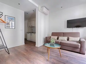 Ein modernes Wohnzimmer mit einer Couch, einem Glastisch, einem an der Wand befestigten Fernseher und einer Küche im Hintergrund.