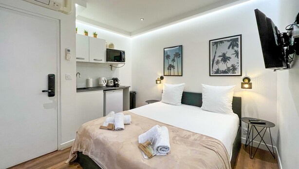 Ein Hotelzimmer mit Bett, einer kleinen Küchenzeile, einem Schreibtisch und einem an der Wand befestigten Fernseher.