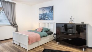Ett sovrum med en säng, ett skrivbord, en stol, ett fönster med gardiner och en tavla med en stadsbild på väggen.