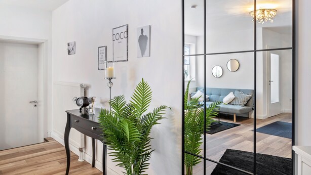 Ein modernes Interieur mit Holzboden, einem schwarzen Konsolentisch, einer Topfpflanze und einer Spiegelwand, die ein Wohnzimmer mit Sofa und einer Chandelier reflektiert.