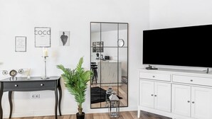 Ein modernes Wohnzimmer mit einem großen Flachbildfernseher, einem spiegelnden Wandbild mit schwarzem Rahmen, einem weißen Schrank, einem kleinen Schreibtisch mit Schubladen und einer Zimmerpflanze.