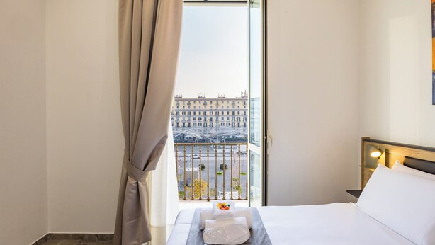 A hotel room with a bed, a nightstand, a window with curtains, and a view of a cityscape. - Sant'Angelo Station (Naples)