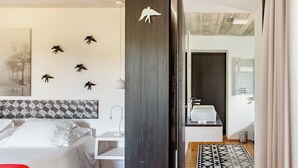 Een slaapkamer met een bed, een nachtkastje, een wandplank, een rode stoel, een kamerbreed tapijt en een badkamer met een wastafel en spiegel.