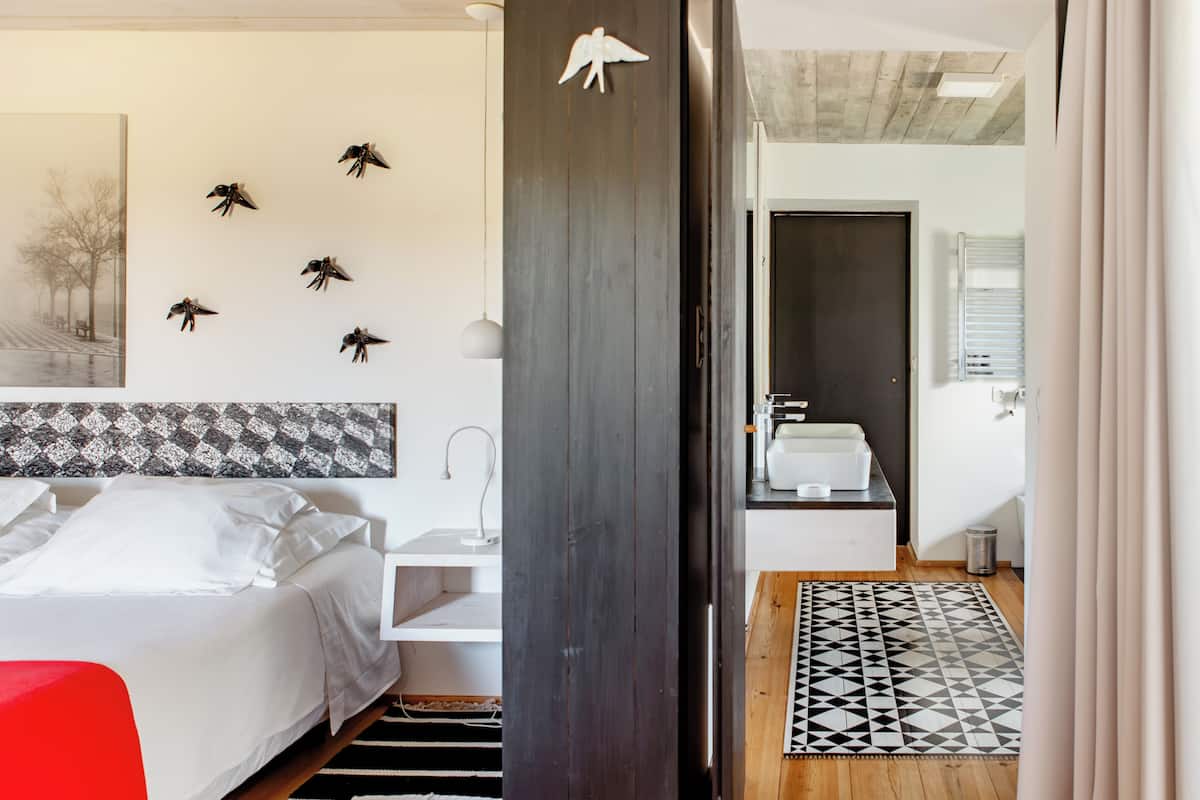 Un dormitorio con cama, mesita de noche, estante en la pared, silla roja, alfombra estampada y baño con lavabo y espejo.