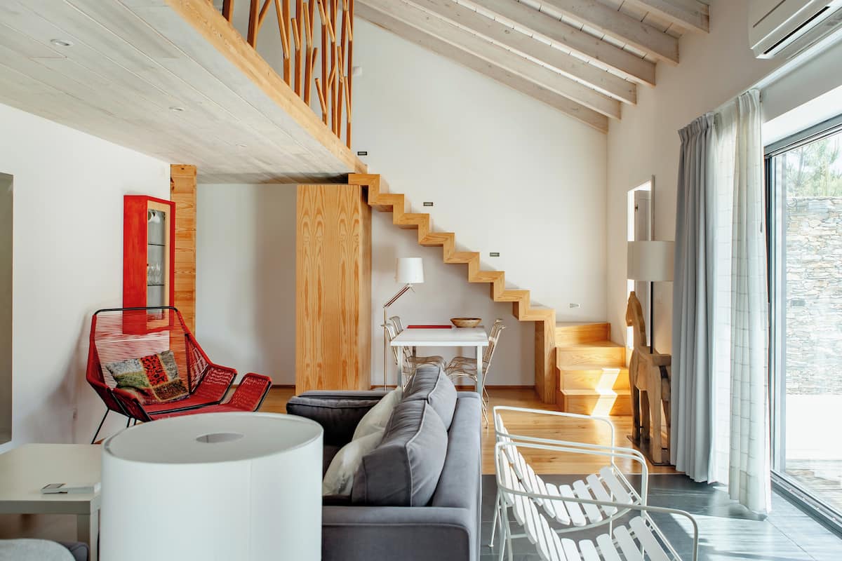 Una sala de estar moderna con un sofá gris, una silla roja, una mesa blanca y una escalera que lleva a un nivel superior.
