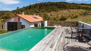 Una piscina con muelle de madera, mobiliario exterior y vistas a una ladera boscosa.