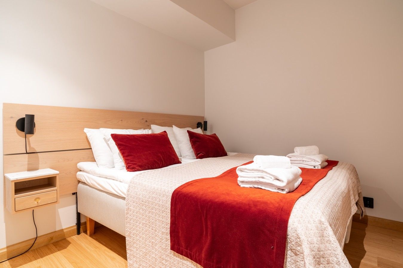 A neatly made bed with white linens and red pillows, a wooden headboard, and a bedside table with a drawer.
