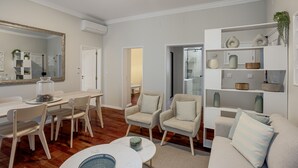 Una sala de estar moderna con comedor, que incluye un espejo grande, muebles blancos y objetos decorativos en estanterías.