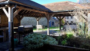 Un'area relax all'aperto coperta, con struttura in legno, un barbecue e un edificio in pietra sullo sfondo.