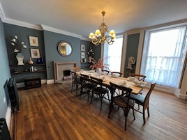 Un comedor con una mesa de madera preparada para seis personas, flanqueada por sillas, una araña y una chimenea.