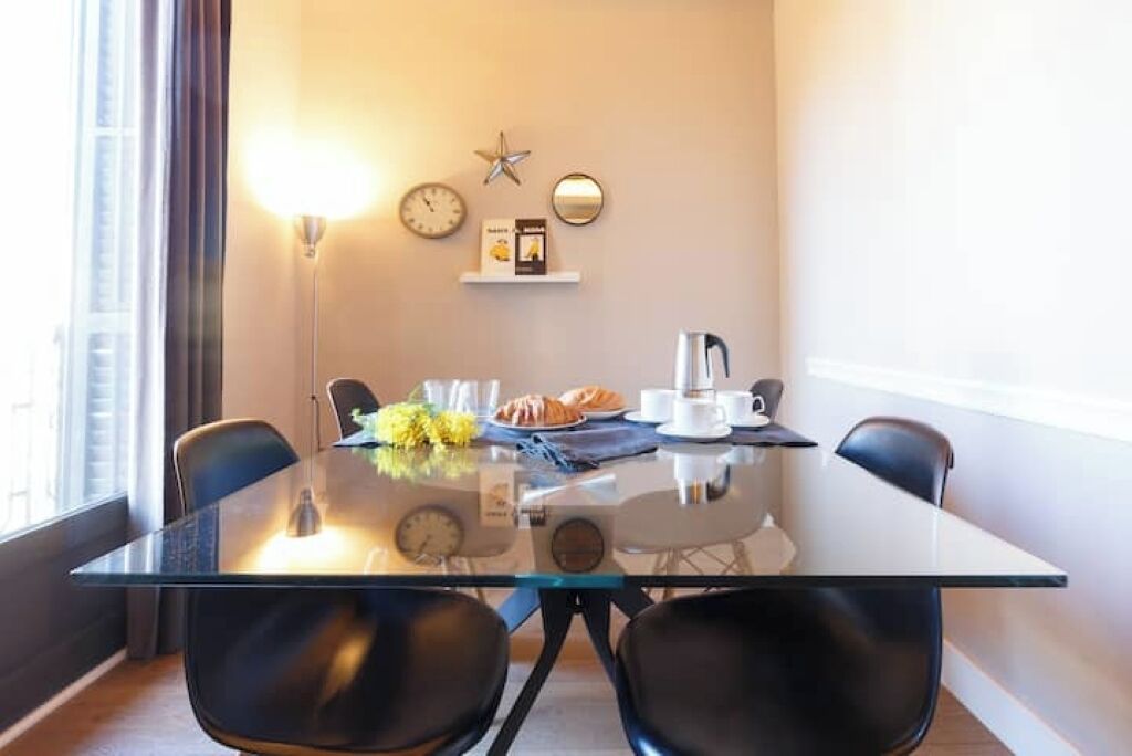 A modern dining area with a glass table, black chairs, a clock, a star decoration, and a shelf with books and a coffee pot.